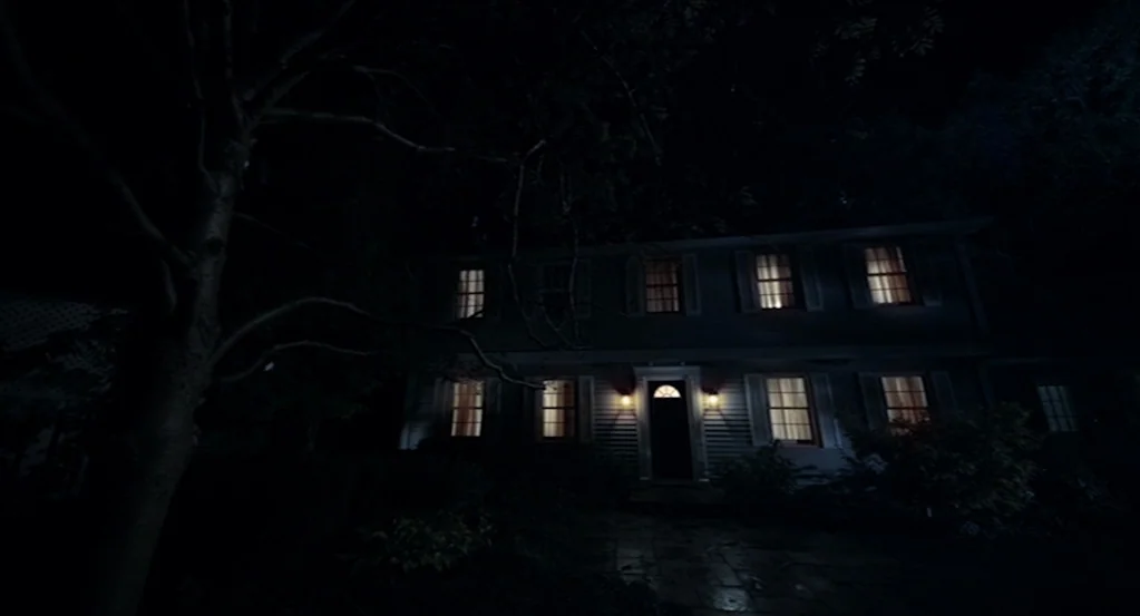 Mama (2013) movie still: wide — A dark two-story suburban house is surrounded by trees at night. Multiple windows emit a soft warm glow against the…