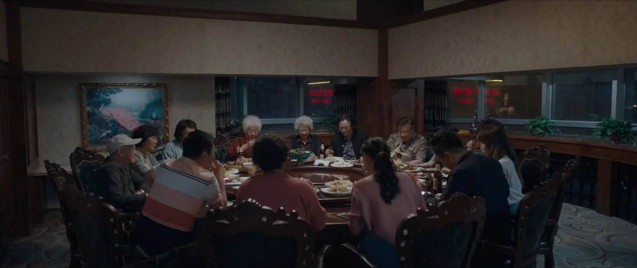 The Farewell (2019) movie still: wide — A group of people sits around a large round dining table in a room. Large windows reveal outdoor neon signs…