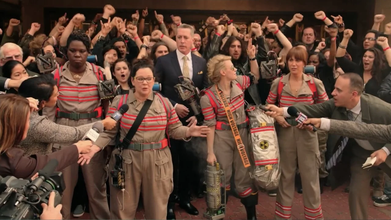Ghostbusters (2016) movie still: medium — A group of people wearing specialized gear stand outdoors before a press gathering. Reporters hold…