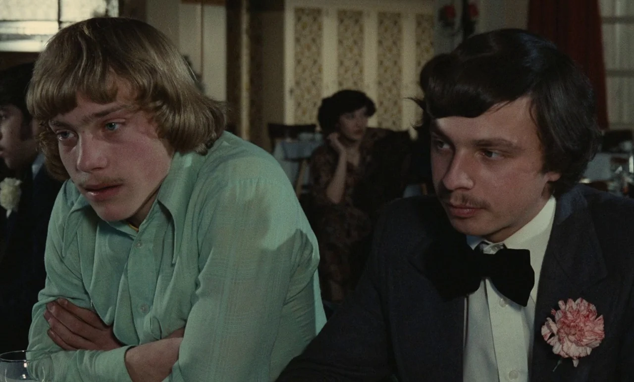 Graduate First (1978) movie still: medium — A banquet hall interior with blurred guests in the background at tables. Two young men sit side-by-side…