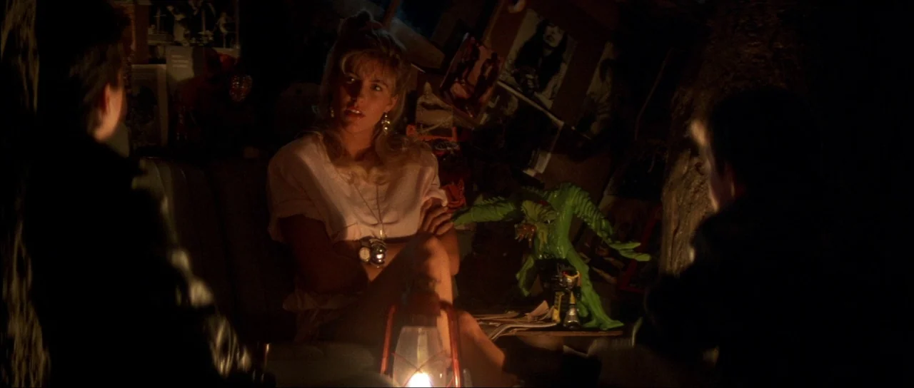 The Monster Squad (1987) movie still: medium — A dark, cluttered attic is filled with posters and toys hanging on the walls. Three young people sit…