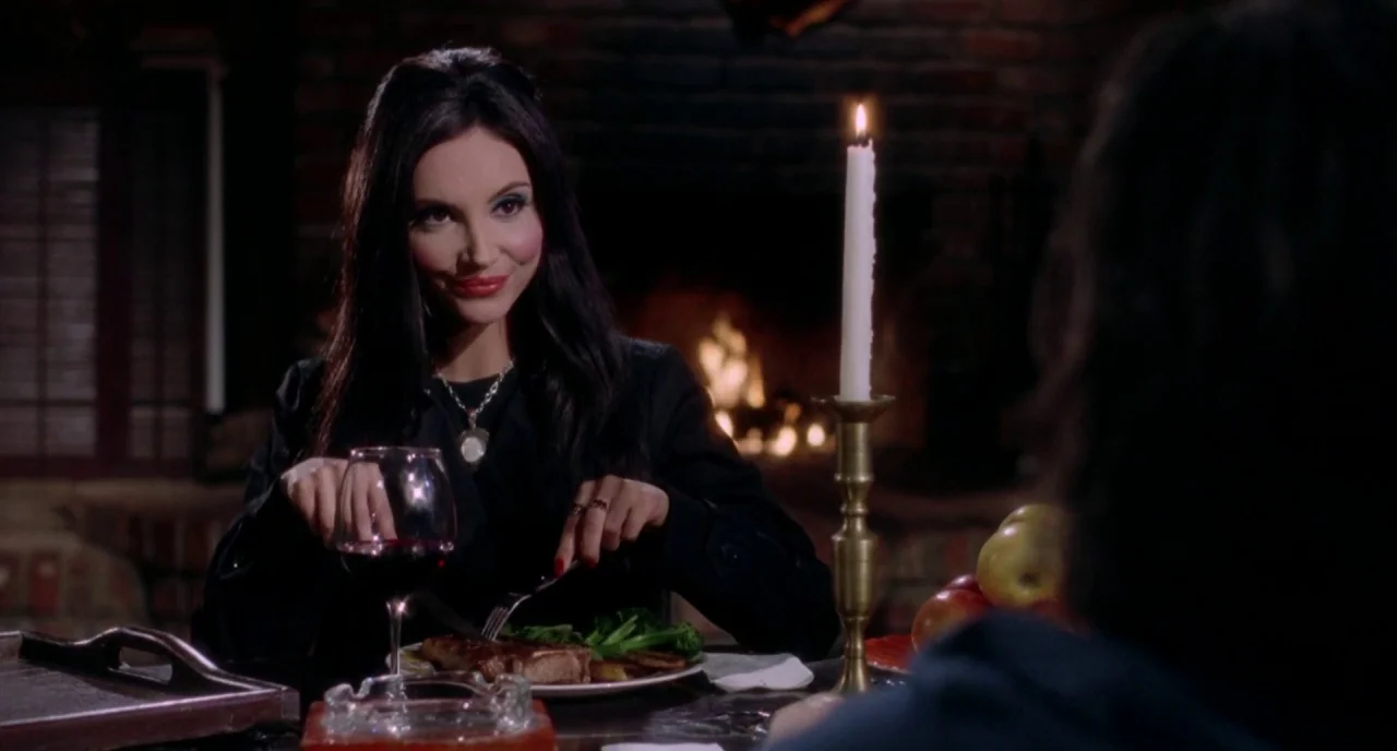 The Love Witch (2016) movie still: medium — A woman sits at a wooden table in front of a flickering fireplace. She holds silverware while eating a…