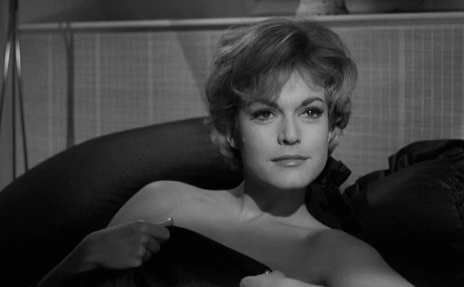 The Criminal (1960) movie still: closeup — A woman lies in a bed with a dark, textured headboard behind her. She looks toward the camera while…