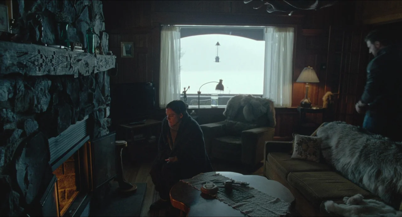 The Lodge (2019) movie still: wide — A rustic cabin living room features stone fireplace masonry, wooden walls, and fur throws. A woman sits on the…
