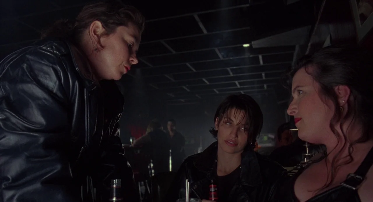 Bound (1996) movie still: medium — A dimly lit room features three women gathered around a table with beverages. They are positioned in a dark,…