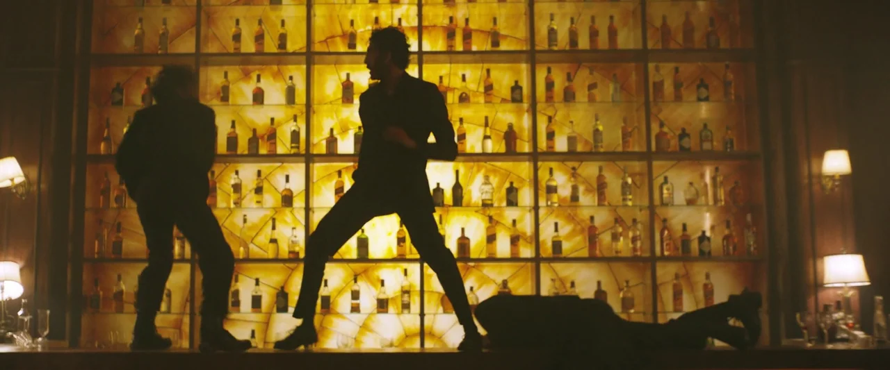 Monkey Man (2024) movie still: wide — A bar interior features an extensive wall of back-lit shelving stocked with liquor bottles. Three men move…