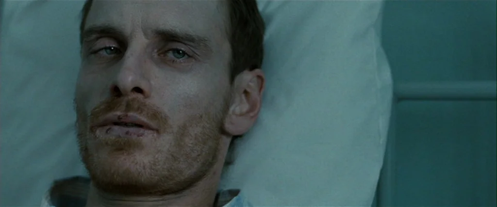 Hunger (2008) movie still: closeup — A man lies on a hospital bed with a white pillow behind his head. The man has facial hair and scabbing around…