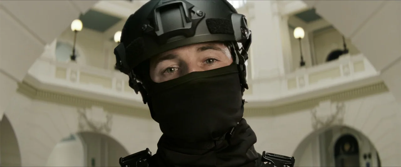 Heads of State (2025) movie still: closeup — A man in tactical gear stands in a bright, multi-level architectural rotunda. He stares directly at the…