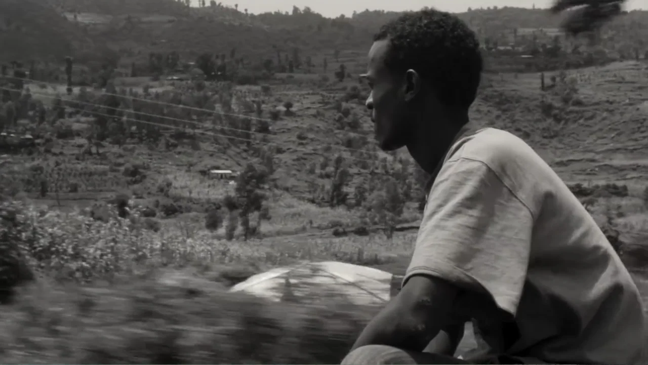 Faya Dayi (2021) movie still: medium — A rural landscape features rolling hills with scattered vegetation and power lines. A young man sits in…
