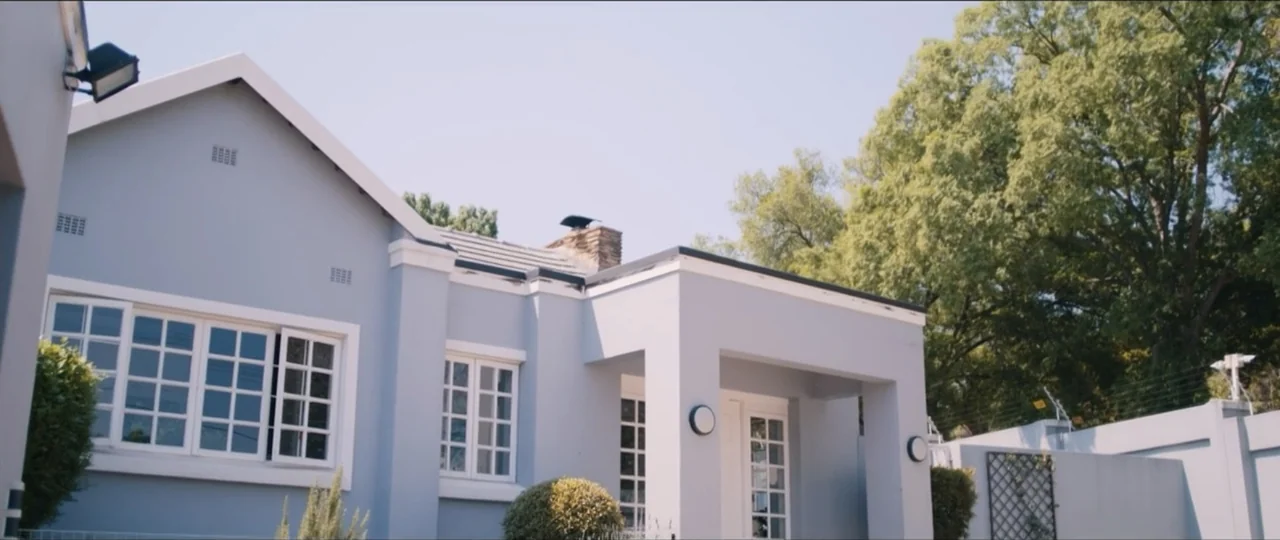 My Girlfriend's Father (2023) movie still: wide — A light blue suburban house with white window frames stands under a bright sky. Lush green trees…