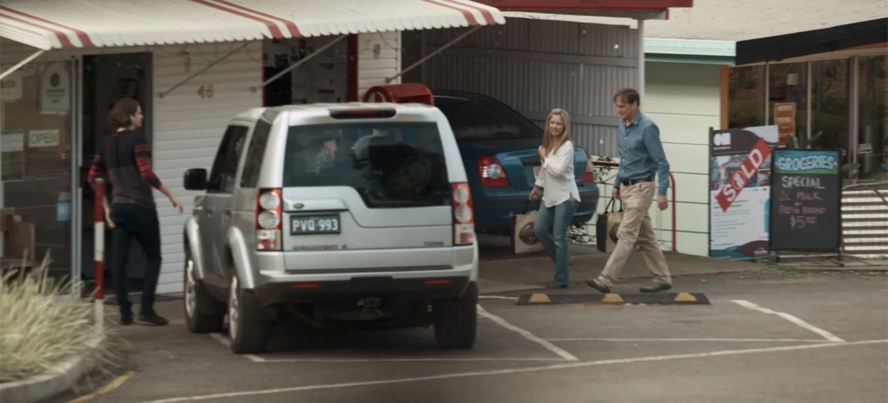 Boar (2018) movie still: wide — A suburban convenience store parking lot features a silver SUV and a small group. Three people walk across the…