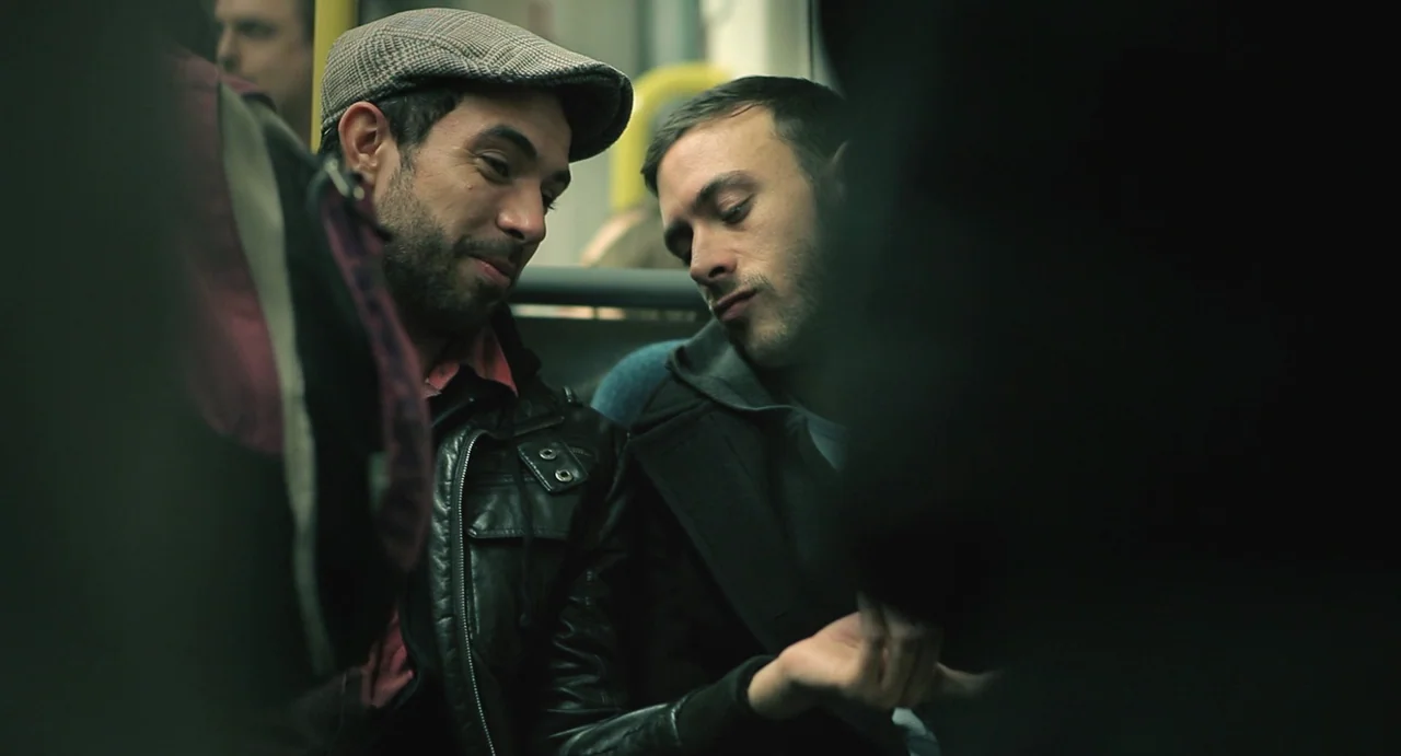 Weekend (2011) movie still: medium — Interior of a modern train carriage with blurred yellow handrails visible in the background. Two young men lean…