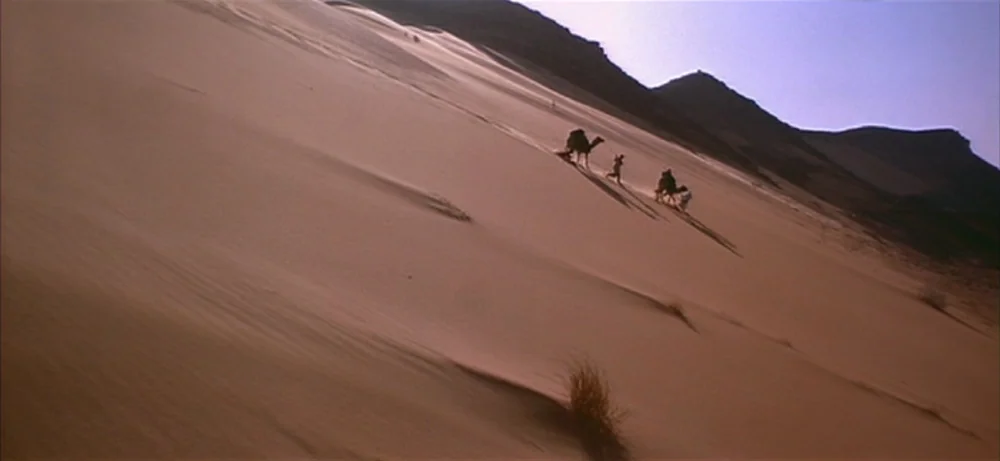 Lawrence Of Arabia movie still: wide — A vast, sun-baked sand dune stretches diagonally across the frame under a clear sky. A small caravan of…