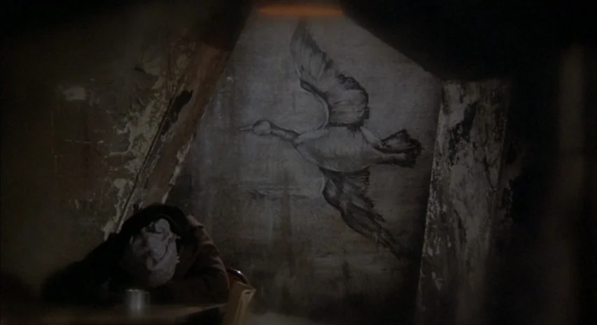 Quintet (1979) movie still: medium — A dimly lit, cramped attic space features peeling paint and uneven wooden walls. A person in a heavy coat sits…