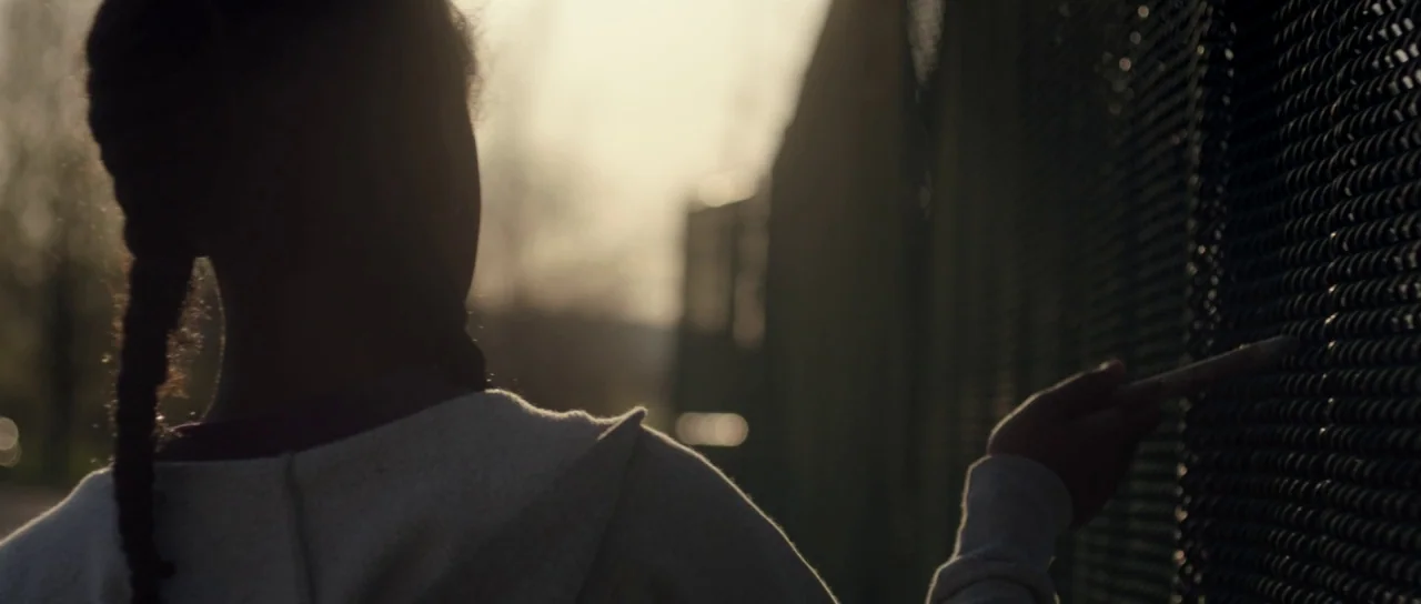 The Fits (2016) movie still: medium — An outdoor area with a tall chain link fence during late afternoon. A young person with braided hair stands…