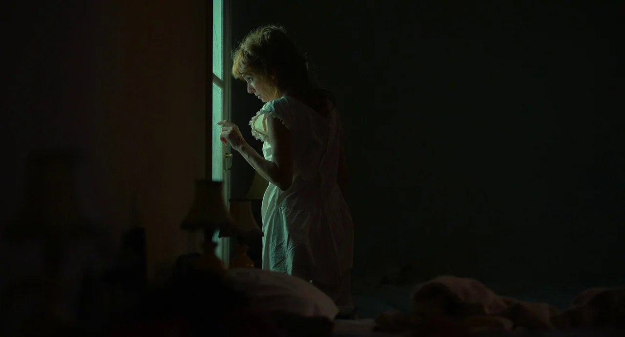 Flux Gourmet (2022) movie still: medium — A dark bedroom interior is illuminated only by the faint light coming from a window. A woman stands near…