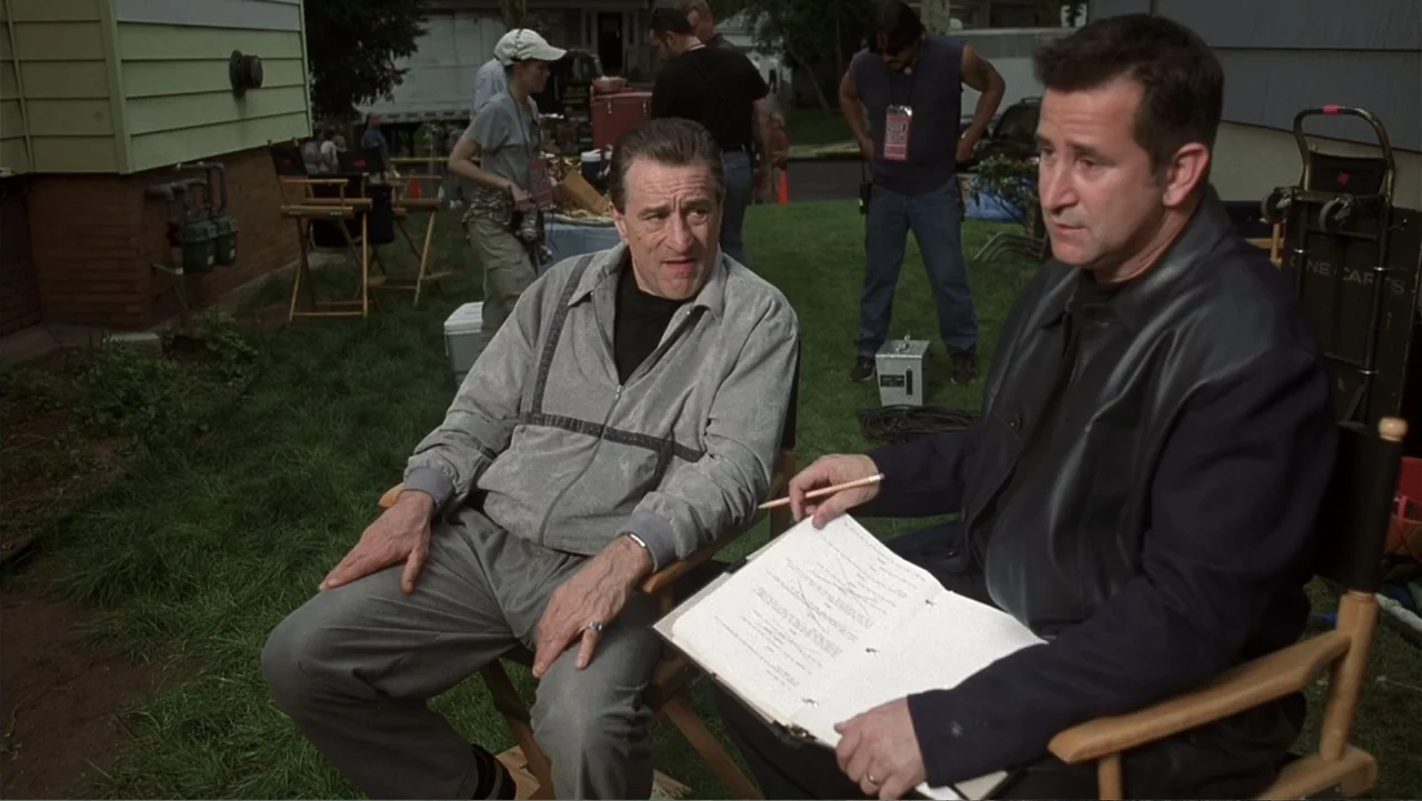 Analyze That (2002) movie still: medium — Two men sit in director chairs on a grassy area adjacent to a building. The man on the right holds an open…