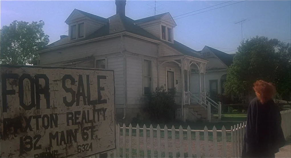 Carrie (1976) movie still: wide — A weather-worn white suburban house features a large For Sale sign in the foreground. A woman with curly hair…