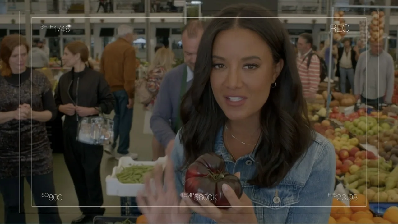 A Pinch of Portugal (2023) movie still: medium — A woman stands in a bright indoor market filled with stalls of produce. She holds a large dark…