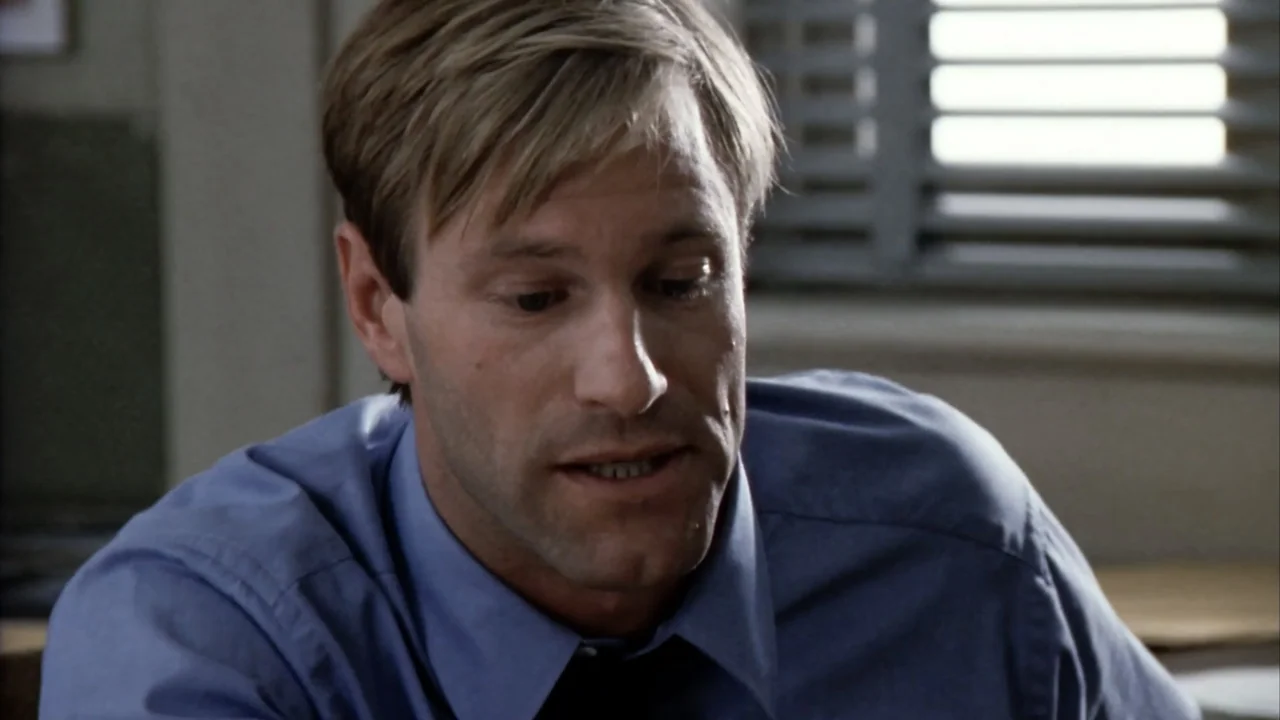 Suspect Zero (2004) movie still: closeup — A man sits in an office space with window blinds visible in the background. The man in a blue dress shirt…