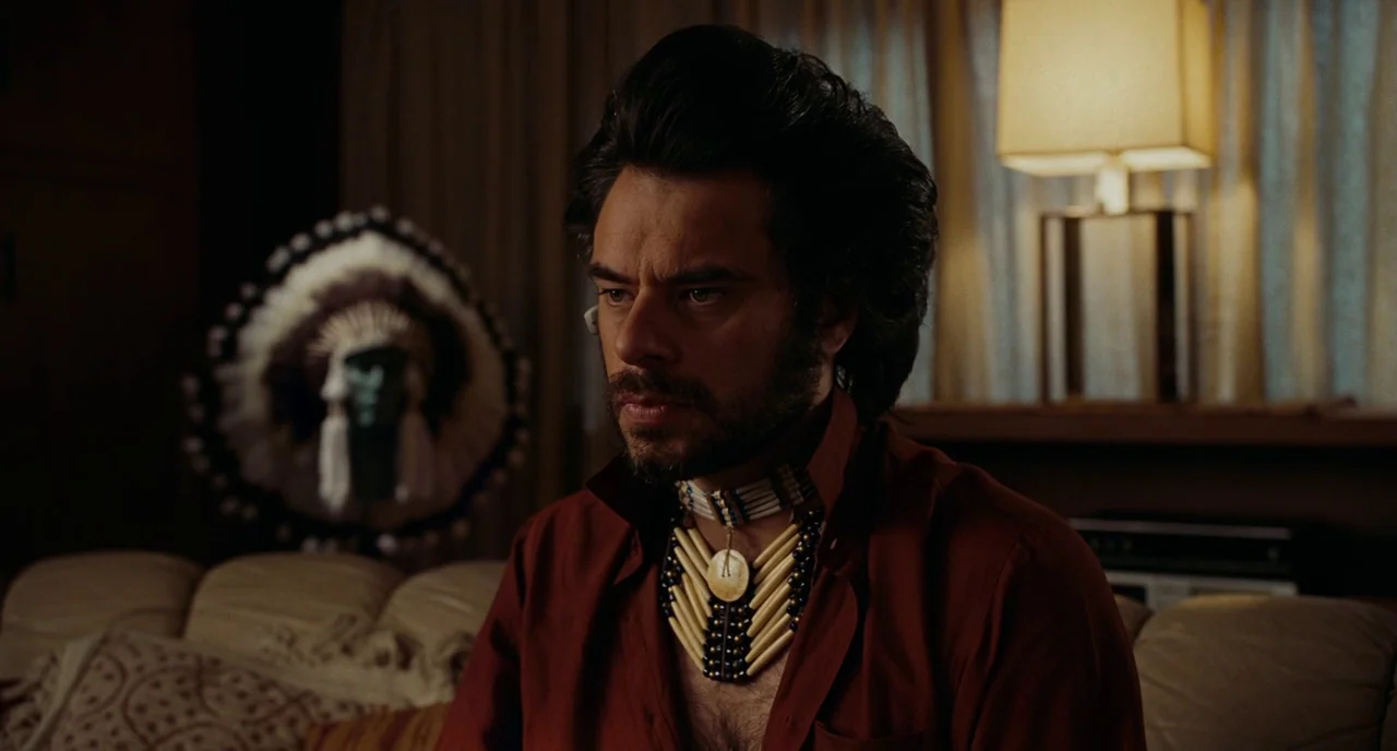Gentlemen Broncos (2009) movie still: medium — A dark living room features a lit lamp and a decorative headdress in back. A man with a beard sits on…