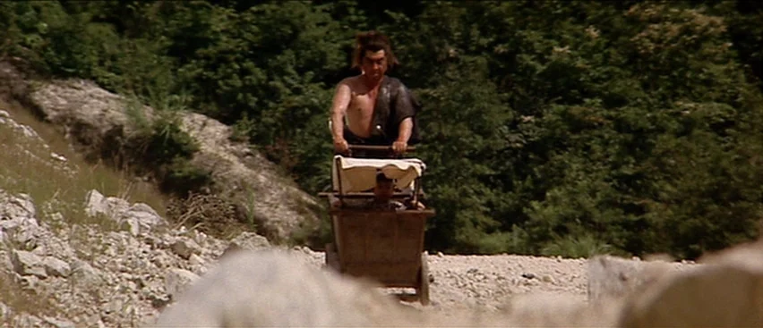 Lone Wolf and Cub: Baby Cart to Hades (1972) movie still: wide — A rugged mountain path covered in gravel is lined with dense green foliage. A man…