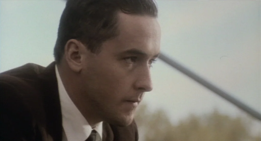 Eight Men Out (1988) movie still: closeup — An outdoor setting features an out-of-focus diagonal railing against a pale sky. A young man in a suit…