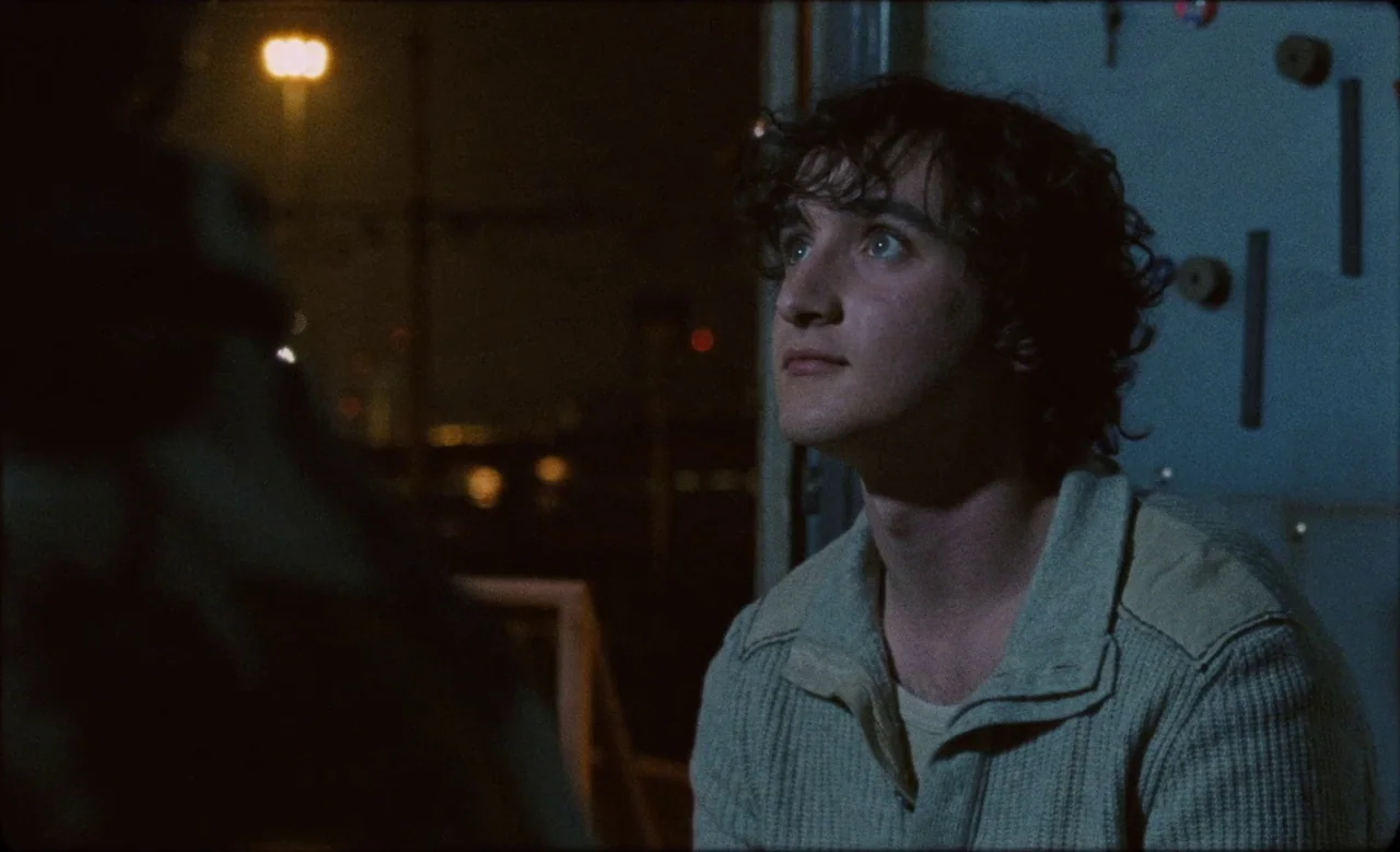 Happy as Lazzaro (2018) movie still: medium — A dark outdoor space at night with distant out-of-focus city lights visible in the background. A young…