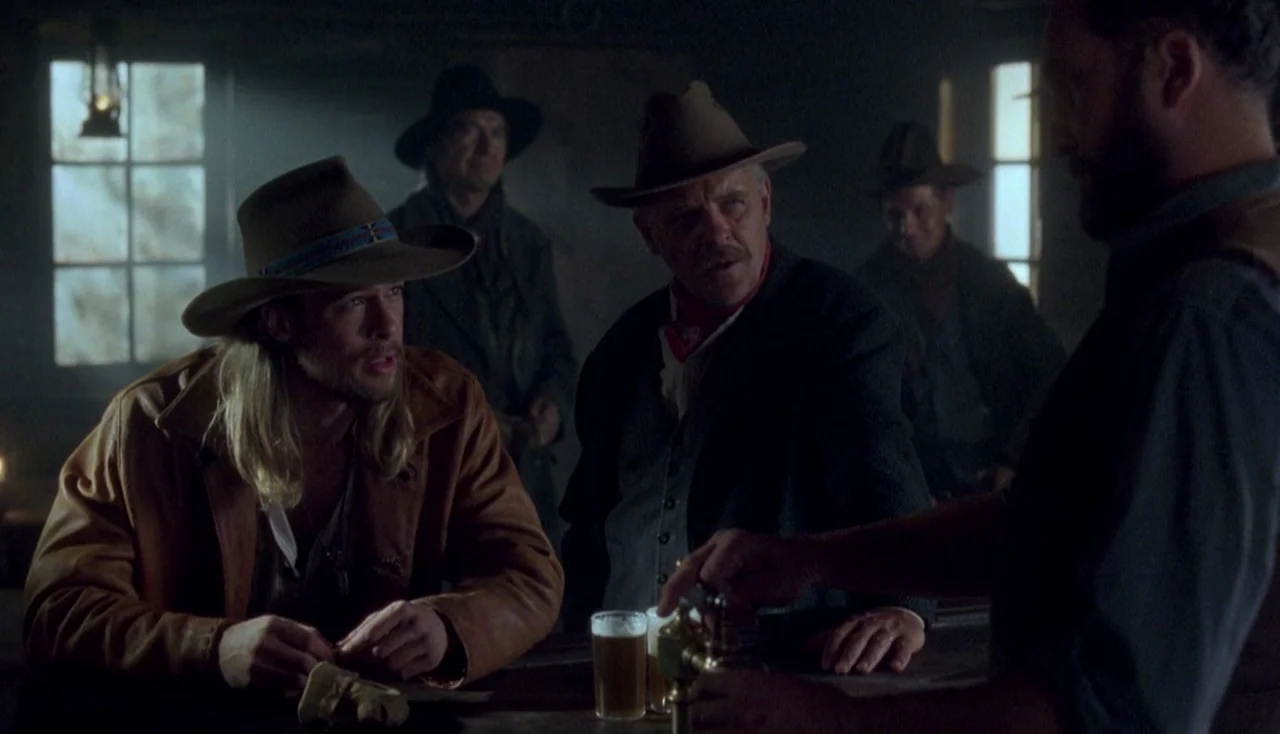 Legends of the Fall (1994) movie still: medium — A dimly lit room features a wooden bar counter and several men. One man with long blonde hair sits…