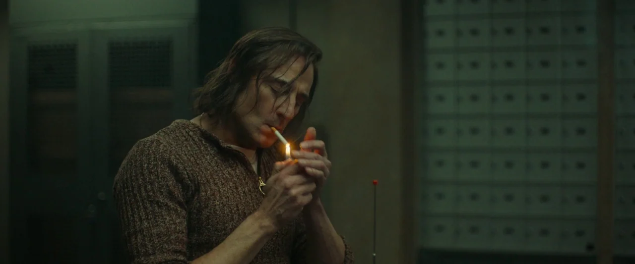 Stockholm (2019) movie still: medium — A dimly lit room features a wall of metallic small storage compartments. A man in a brown sweater lights a…