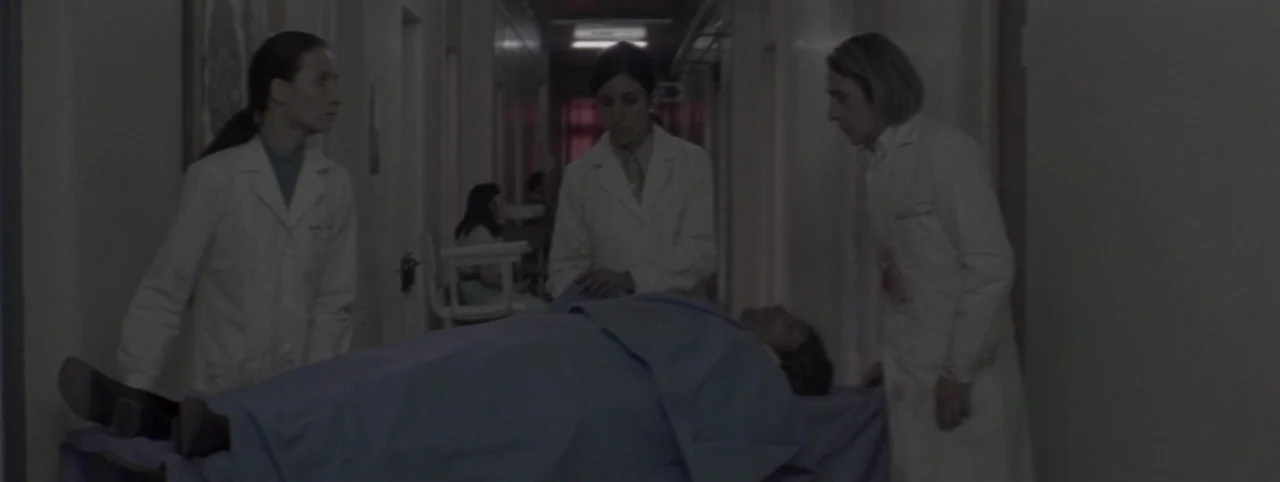 Post Mortem (2010) movie still: medium — A dimly lit hospital corridor with multiple doors lining the walls and a gurney. Three medical staff…