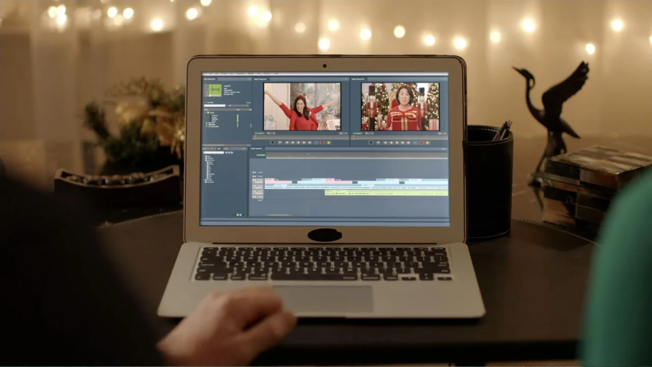 Loving Christmas (2022) movie still: medium — A person sits at a dark desk using a silver laptop computer. The screen displays video editing…