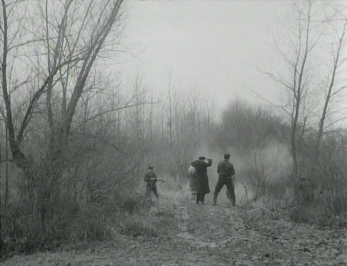 Les Carabiniers (1963) movie still: wide — A desolate dirt path winds through a collection of barren leafless trees and brush. Several people stand…