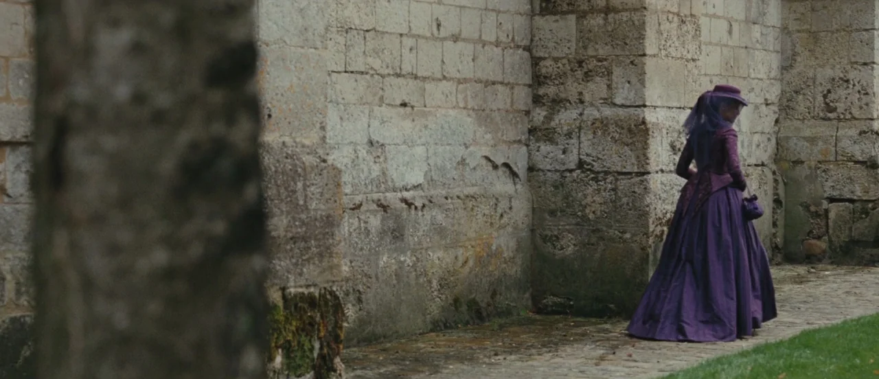 Madame Bovary movie still: wide — A woman in a period purple dress walks beside an ancient stone church wall. She moves along a narrow cobblestone…