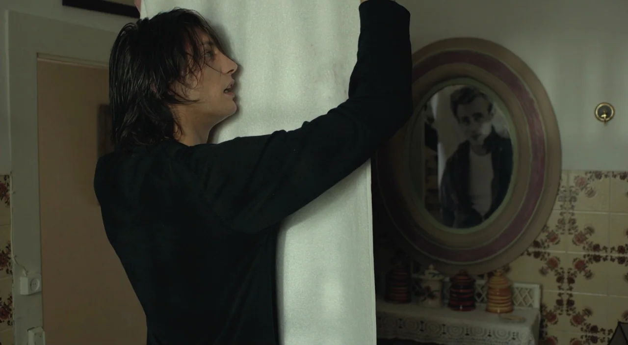 Cosmos (2015) movie still: medium — An interior space features floral-patterned wallpaper and an oval-framed mirror hanging on the wall. A man with…