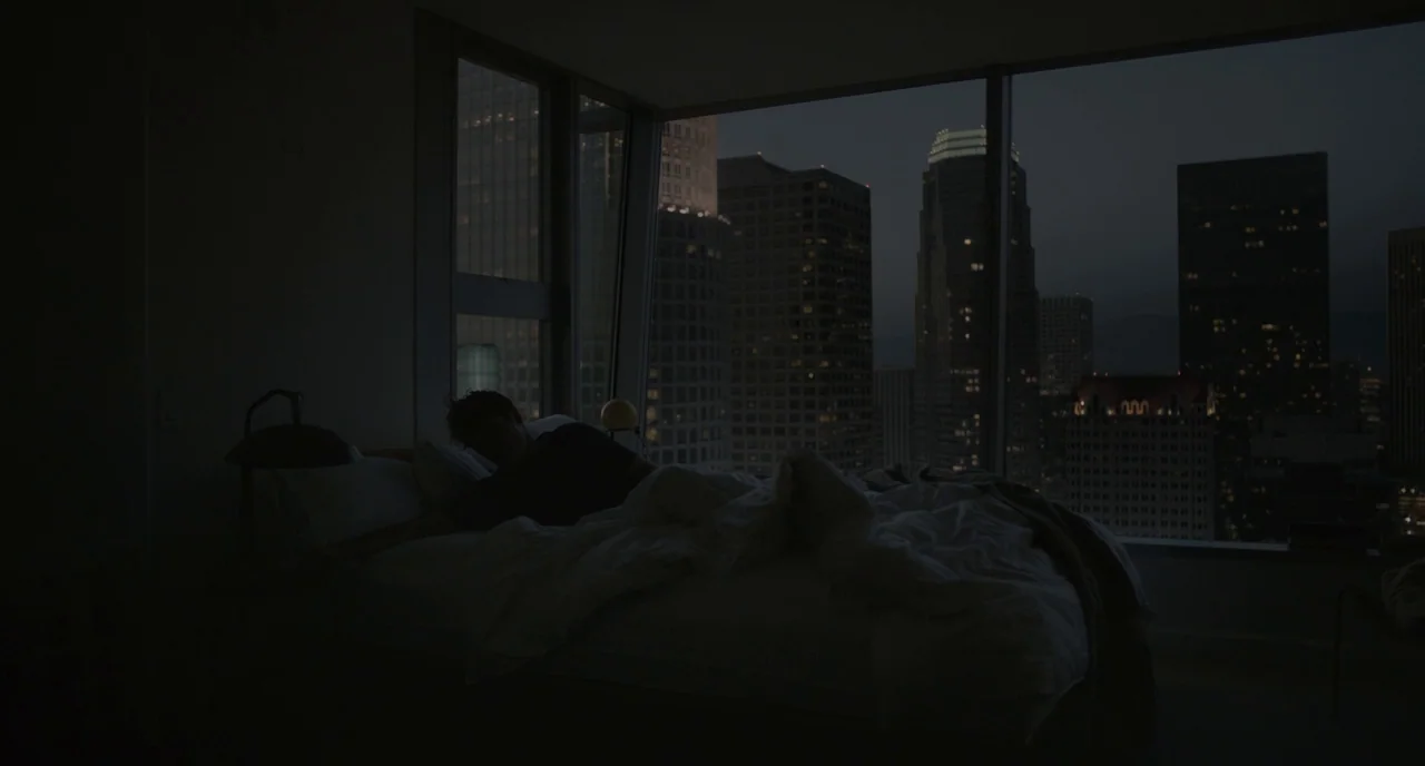 Her (2013) movie still: wide — A high-rise bedroom at night overlooks a dense city skyline of lit skyscrapers. A man lies asleep in a bed with white…