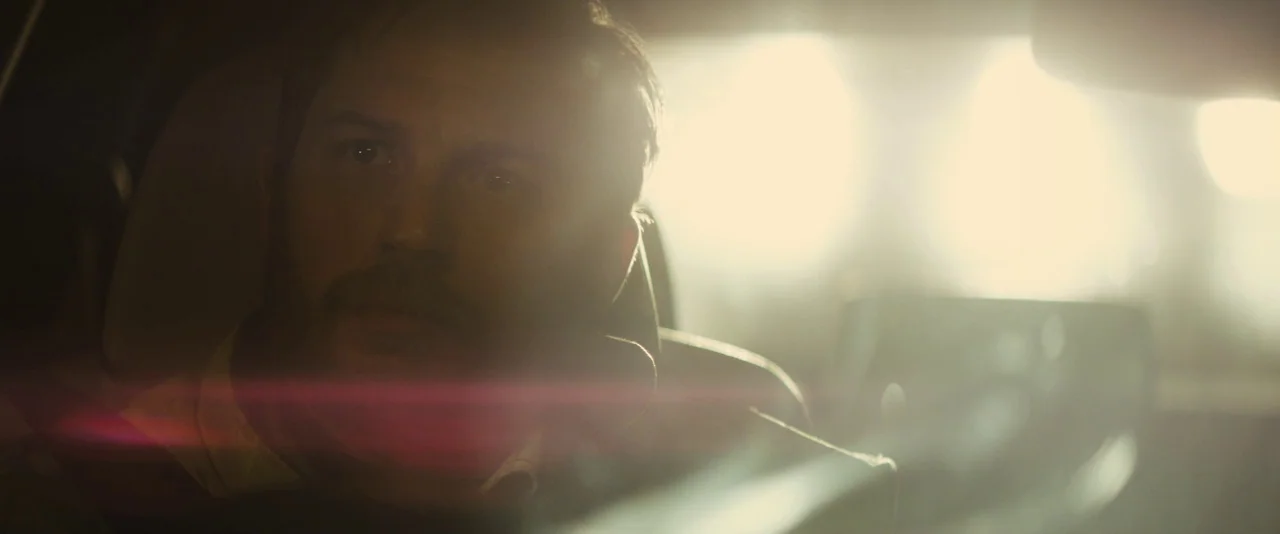 Locke (2013) movie still: closeup — A man drives a car at night with intense light blinding the field of view. The man focuses on the road ahead…