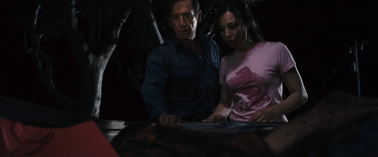 The Loved Ones (2009) movie still: medium — An outdoor night setting featuring a dark area near a tree line. Two people lean over the edge of a…