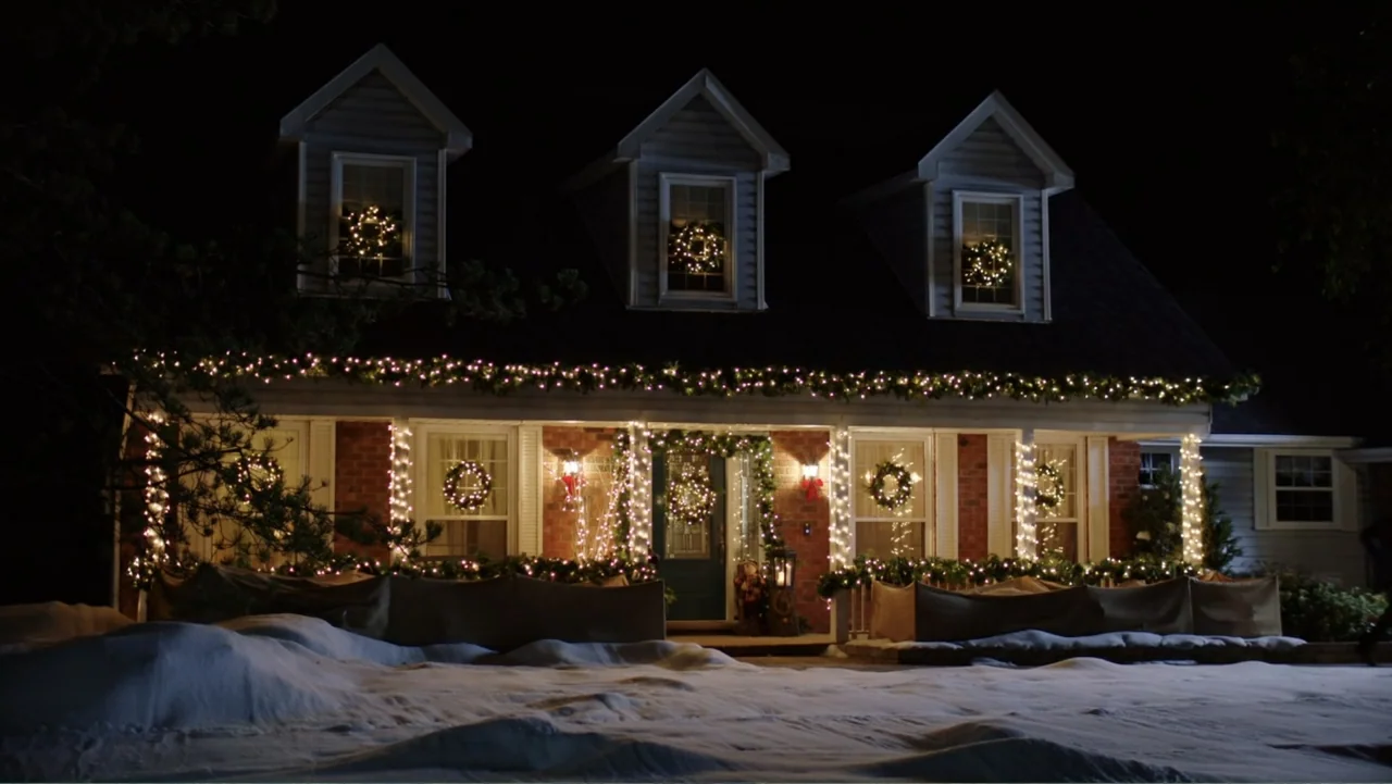 Ghosts of Christmas Past (2021) movie still: wide — A two-story suburban brick house at night is covered in snow and Christmas lights. Decorated…