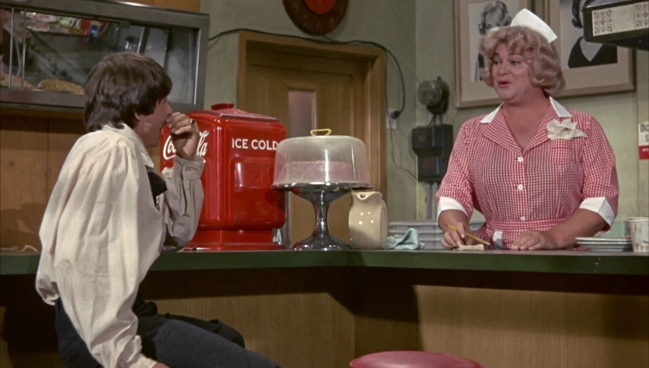 Head (1968) movie still: medium — A retro 1960s diner with wood paneling and a large red soda cooler. A young man sits at the counter while a woman…