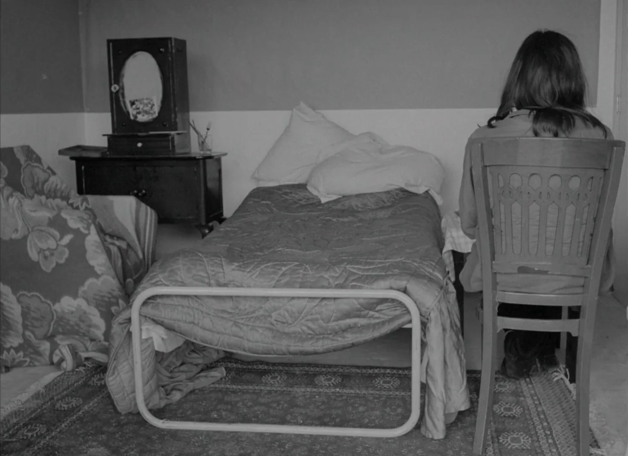 Je Tu Il Elle (1974) movie still: medium — A sparse room contains a metal-framed bed and a small vanity table. A woman sits on a wooden chair,…