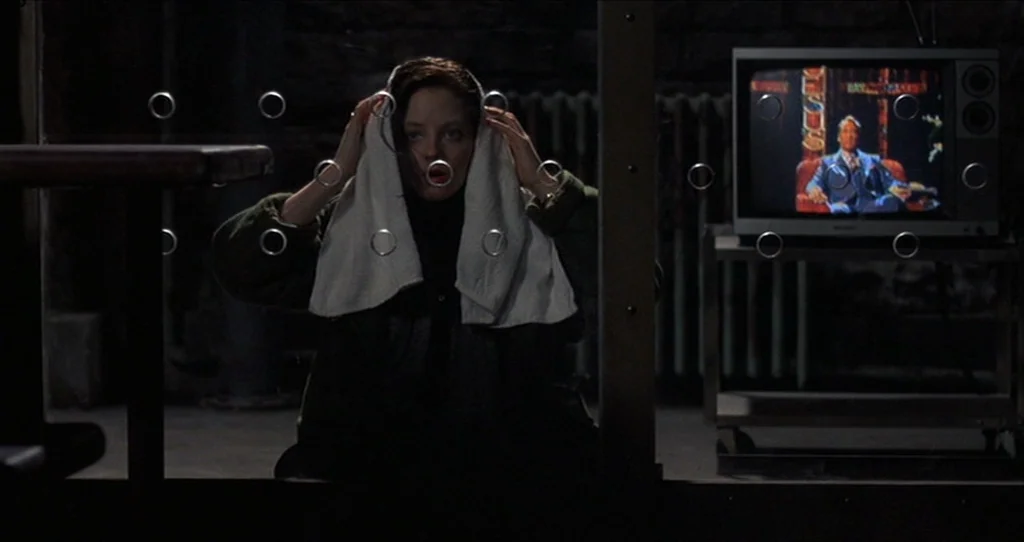 The Silence Of The Lambs (1991) movie still: medium — A dark, spartan observation space features a glass partition with metal grommets. A woman sits…