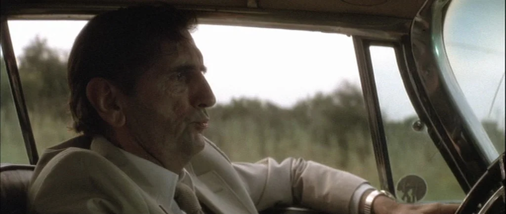 Wild At Heart (1990) movie still: medium — The interior of a vintage car with chrome window frame details. An older man sits in the driver's seat…