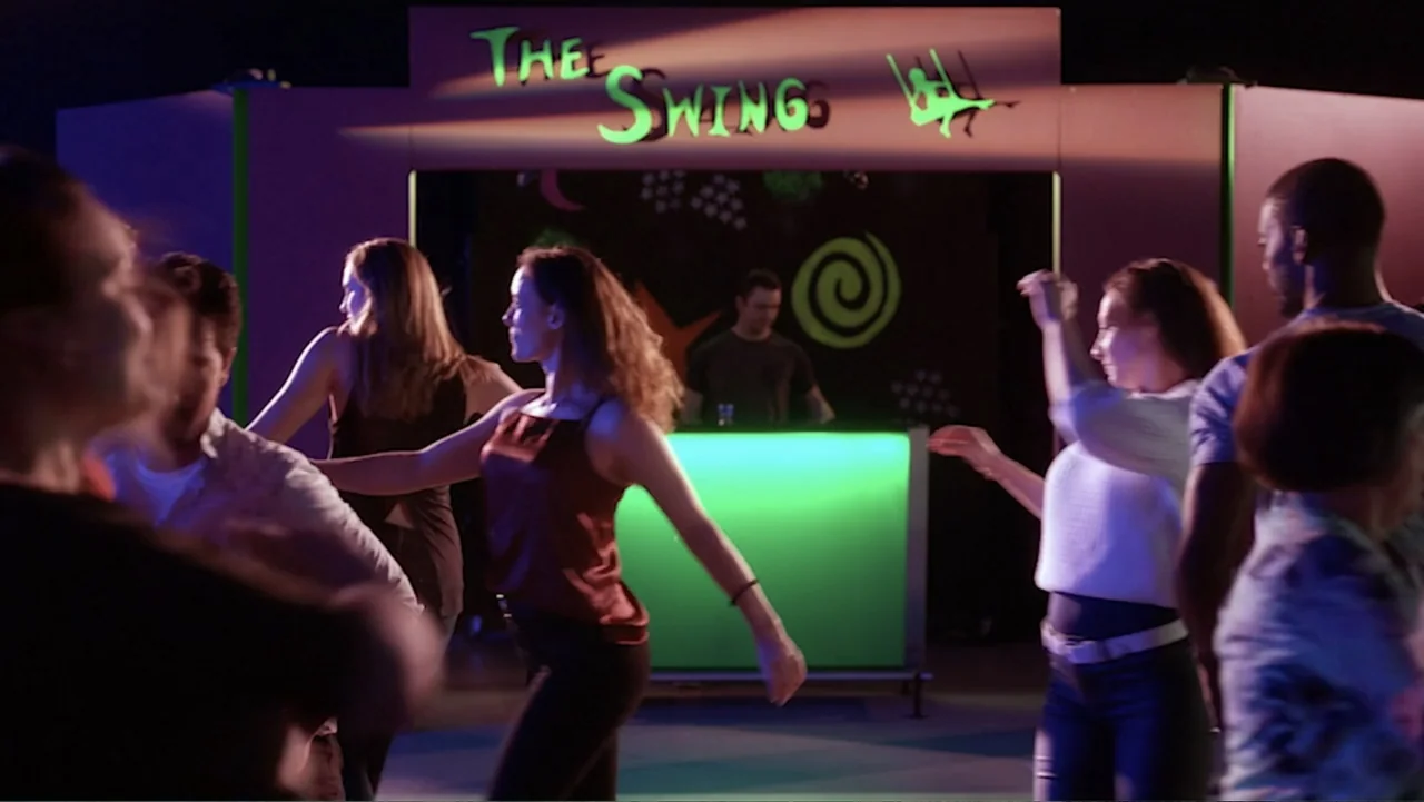 Ask Me to Dance (2022) movie still: medium — A dimly lit dance floor features a sign reading The Swings above a DJ. Several young people move…