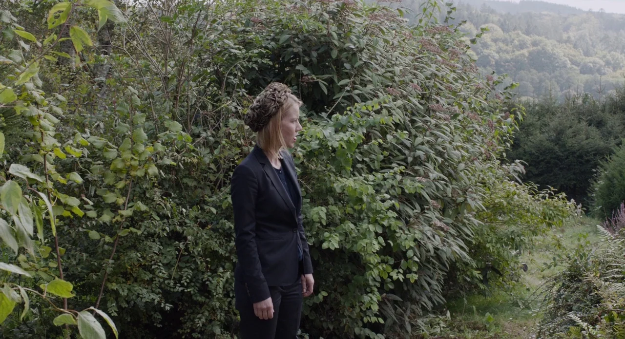 Toni Erdmann (2016) movie still: medium — A woman stands in front of a large, dense, dark green hedge outdoors. The background reveals a blurred,…