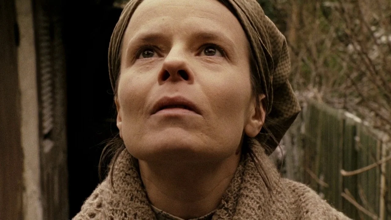 Everlasting Moments (2008) movie still: closeup — A woman stands in a rural outdoor setting with a blurred wooden structure nearby. She looks toward…