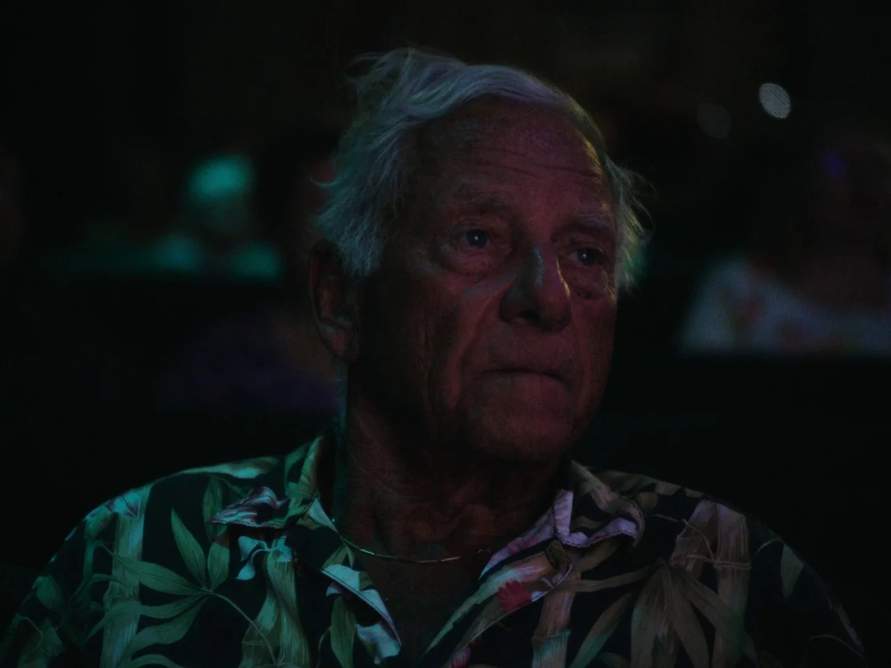 Some Kind Of Heaven (2020) movie still: closeup — An elderly man sits in a darkened space illuminated by light from a screen. He is facing away from…