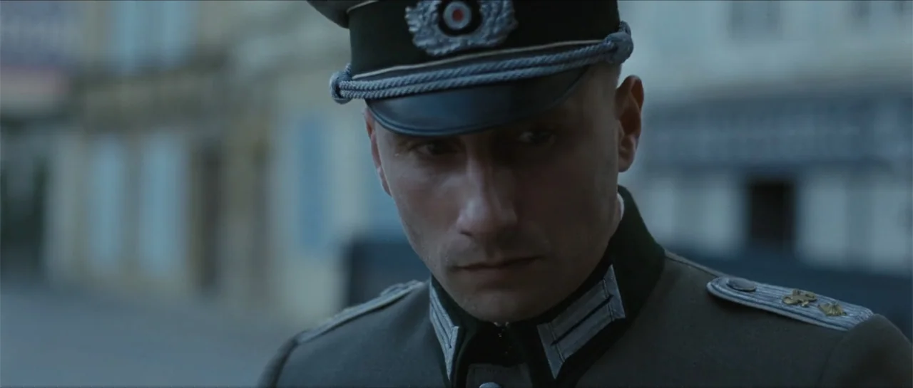 Suite Française (2015) movie still: closeup — An outdoor urban location with blurred building facades in the background. A man wearing a mid-century…