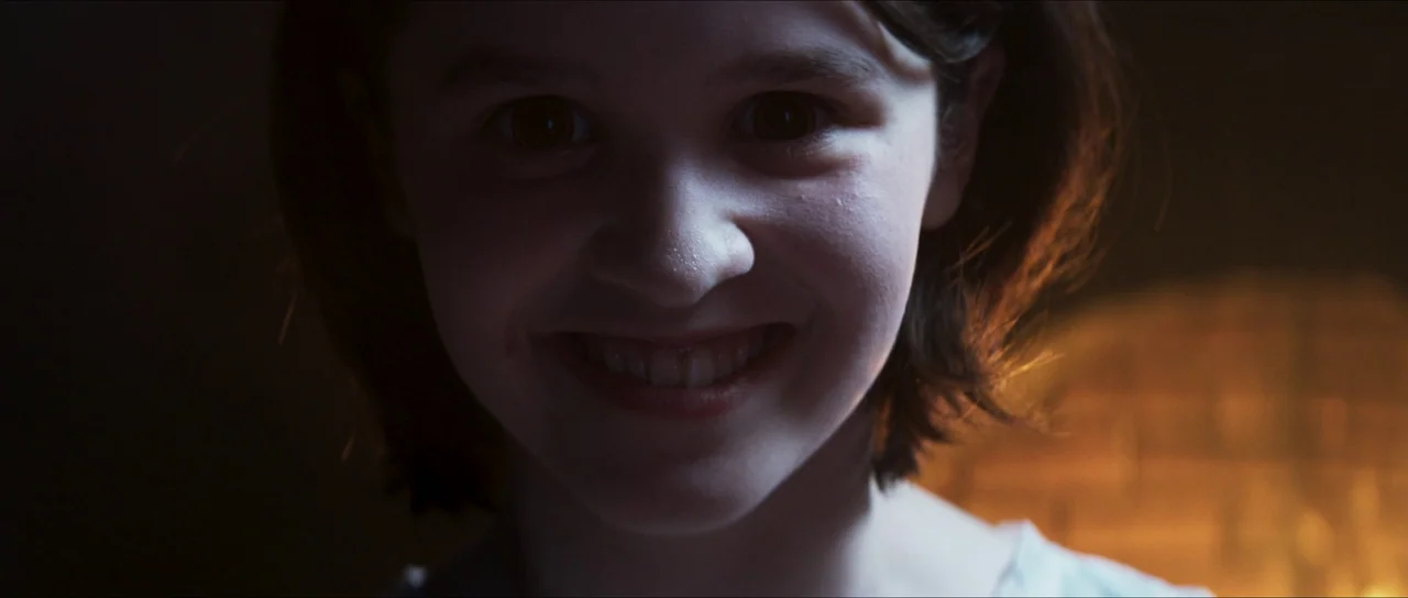 The Sacrifice Game (2023) movie still: closeup — A dark indoor setting features a child partially lit by a warm, side-directional light source. The…