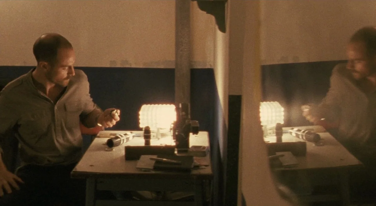 Patagonia (2010) movie still: medium — A small room features a white table against a wall painted with a blue stripe. A bald man sits at the table…